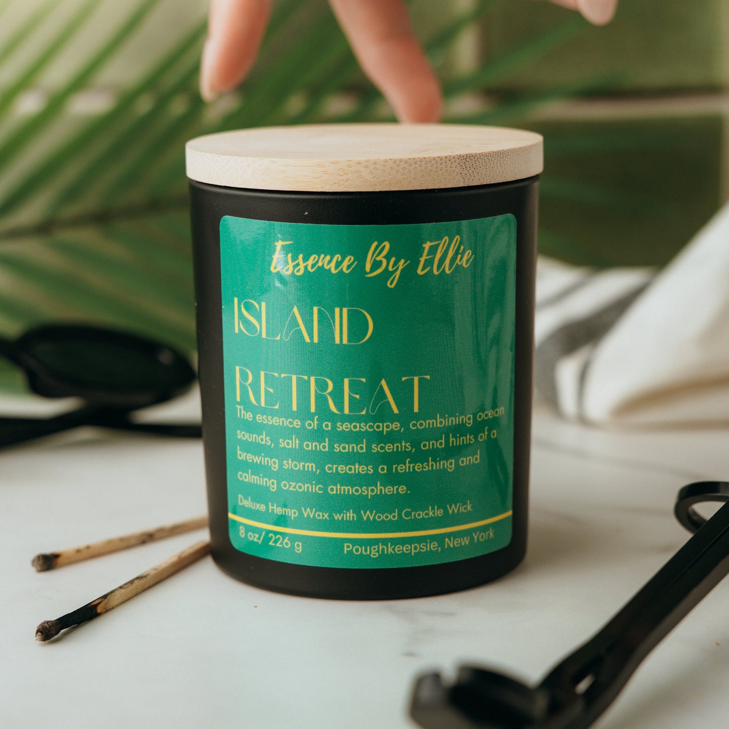 Candle labeled 'Island Retreat' by Essence By Ellie on a surface with a palm leaf, wick trimmer, and two prior lit match sticks in blurred background.
