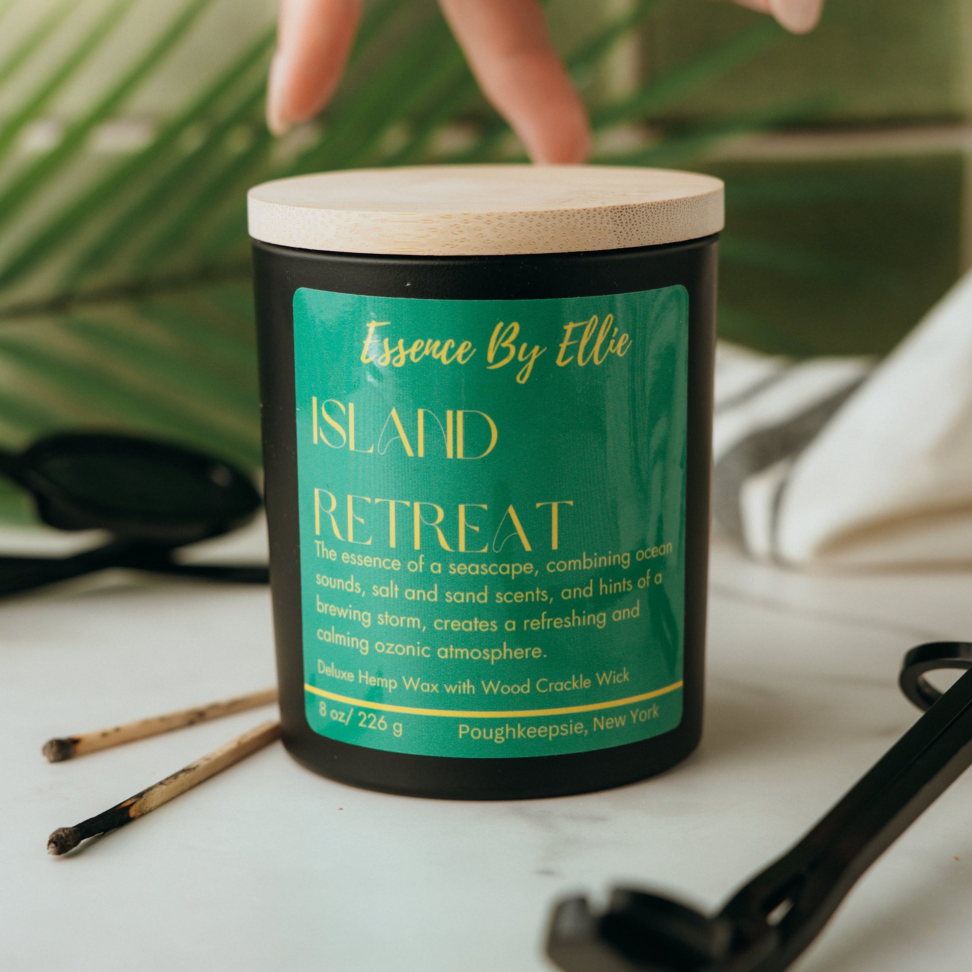 Candle labeled 'Island Retreat' by Essence By Ellie on a surface with a palm leaf, wick trimmer, and two prior lit match sticks in blurred background.