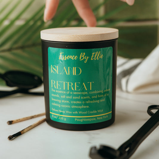 Candle labeled 'Island Retreat' by Essence By Ellie on a surface with a palm leaf, wick trimmer, and two prior lit match sticks in blurred background.