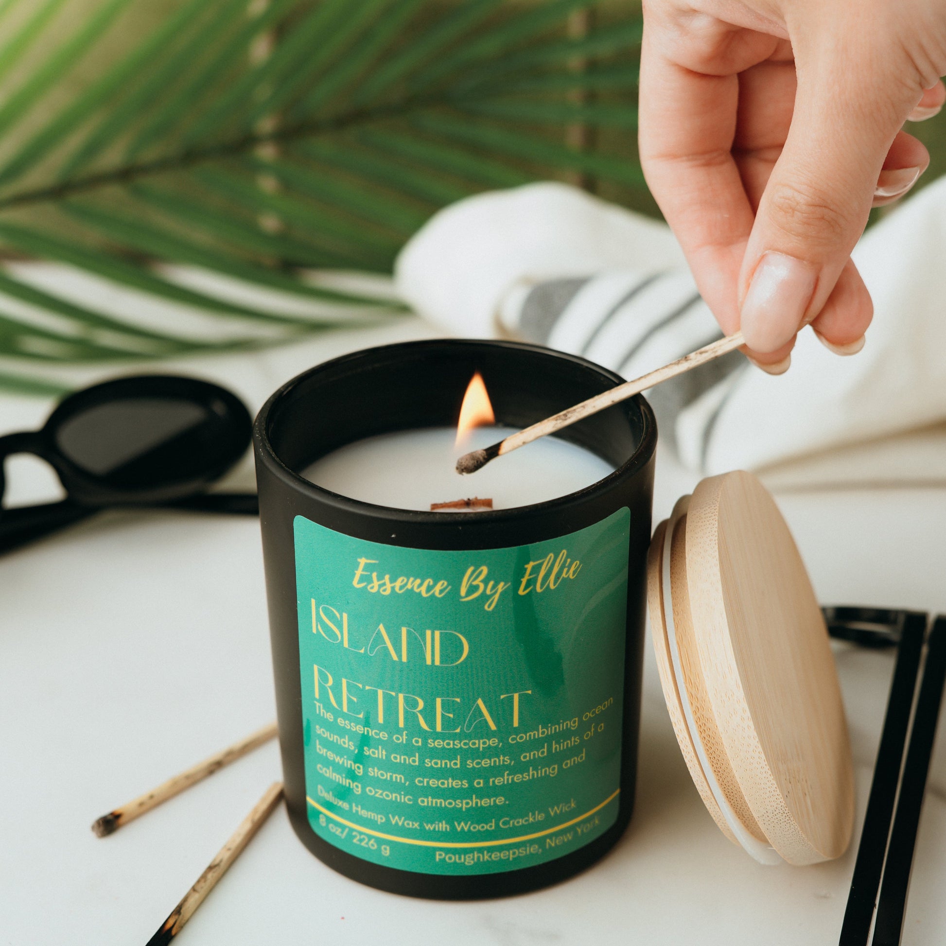 Crackling Wick Candle labeled 'Island Retreat' being lit with a match, surrounded by sunglasses and a plant in background.