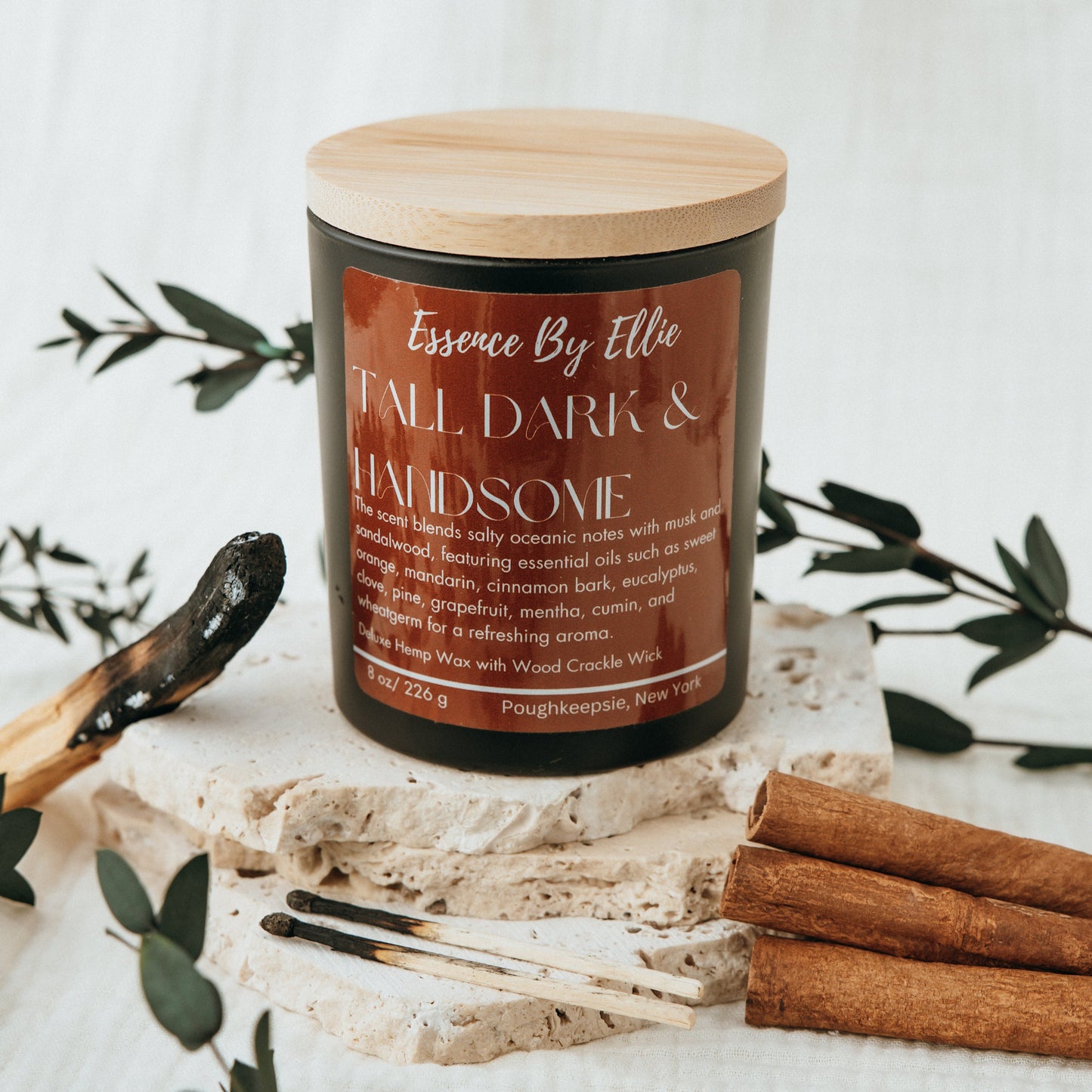 Crackling wick Candle poured in a black vessel with bamboo lid picture is surround by the essence of palo santo cinnamon eucalyptus plants with a soft white background