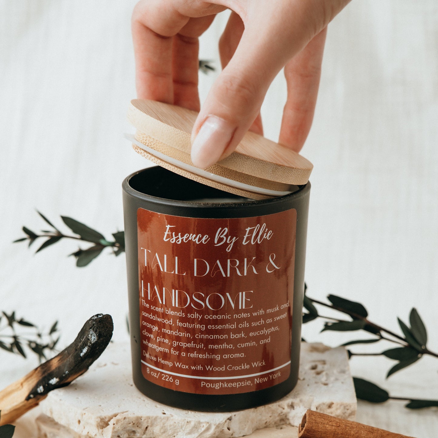 Crackling wick Candle poured in a black vessel with bamboo lid picture of a person's hand opening the lid of the vessel.  The candle is surround by the essence of palo santo cinnamon eucalyptus plants with a soft white background