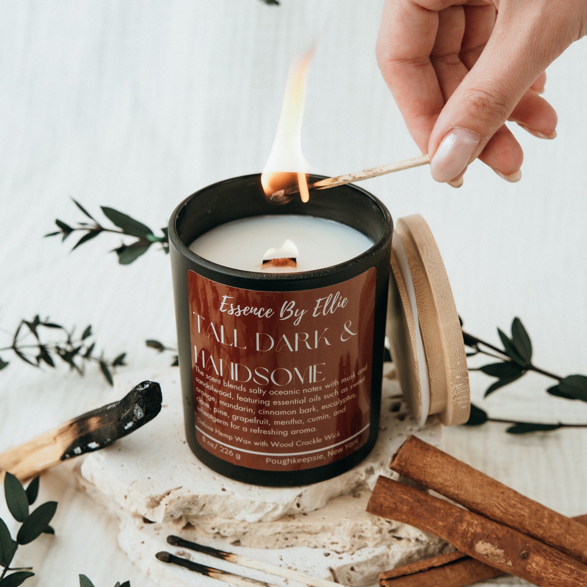 Crackling wick Candle poured in a black vessel with bamboo lid picture of a lit match and candle surround by the essence of sage cinnamon eucalyptus plants with a soft white background
