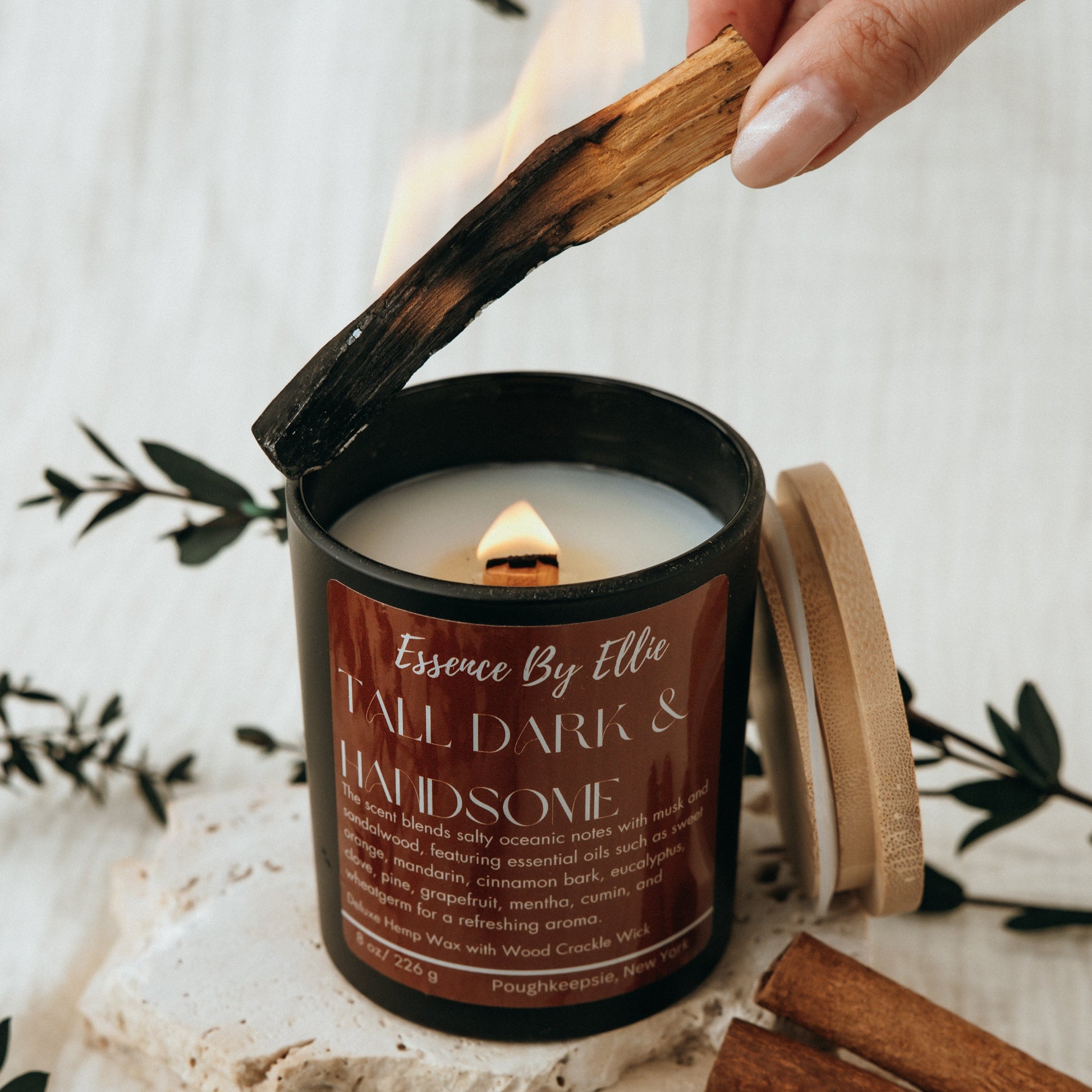 Crackling wick Candle poured in a black vessel with bamboo lid picture of a lit palo santo and candle surround by the essence of palo santo cinnamon eucalyptus plants with a soft white background